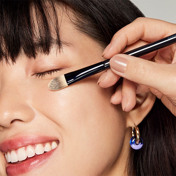 An image of a woman having her undereye area brightened with concealer on a brush